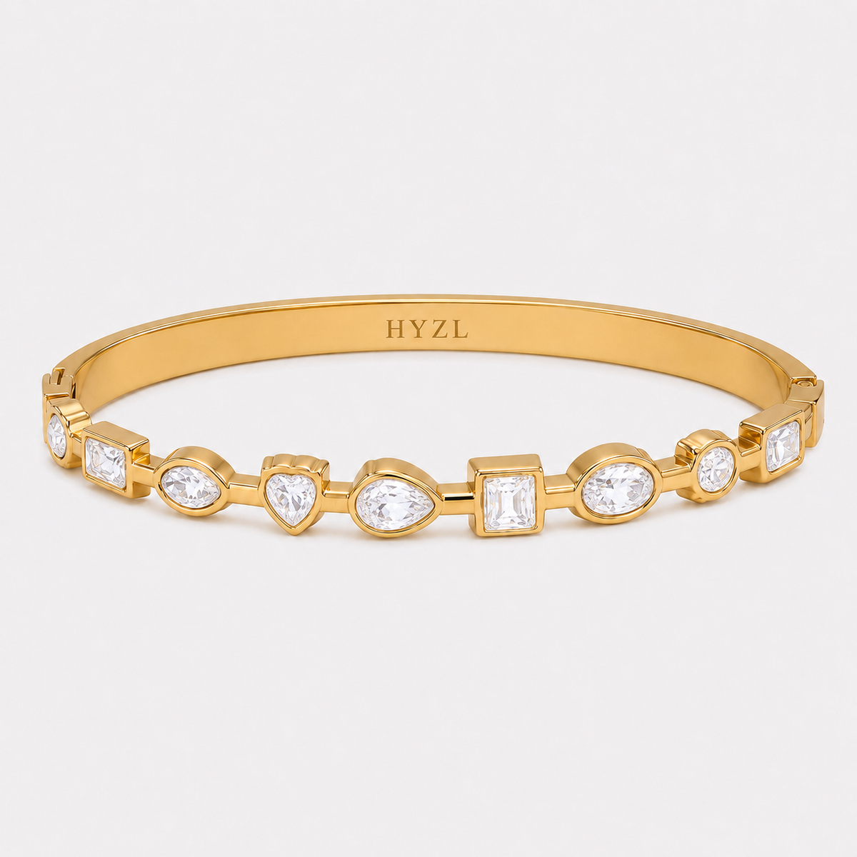 A gold hinged bangle bracelet with a row of clear gemstones in heart, pear, and rectangular shapes, featuring the brand name 'HYZL' engraved inside the band, set against a light gray background.