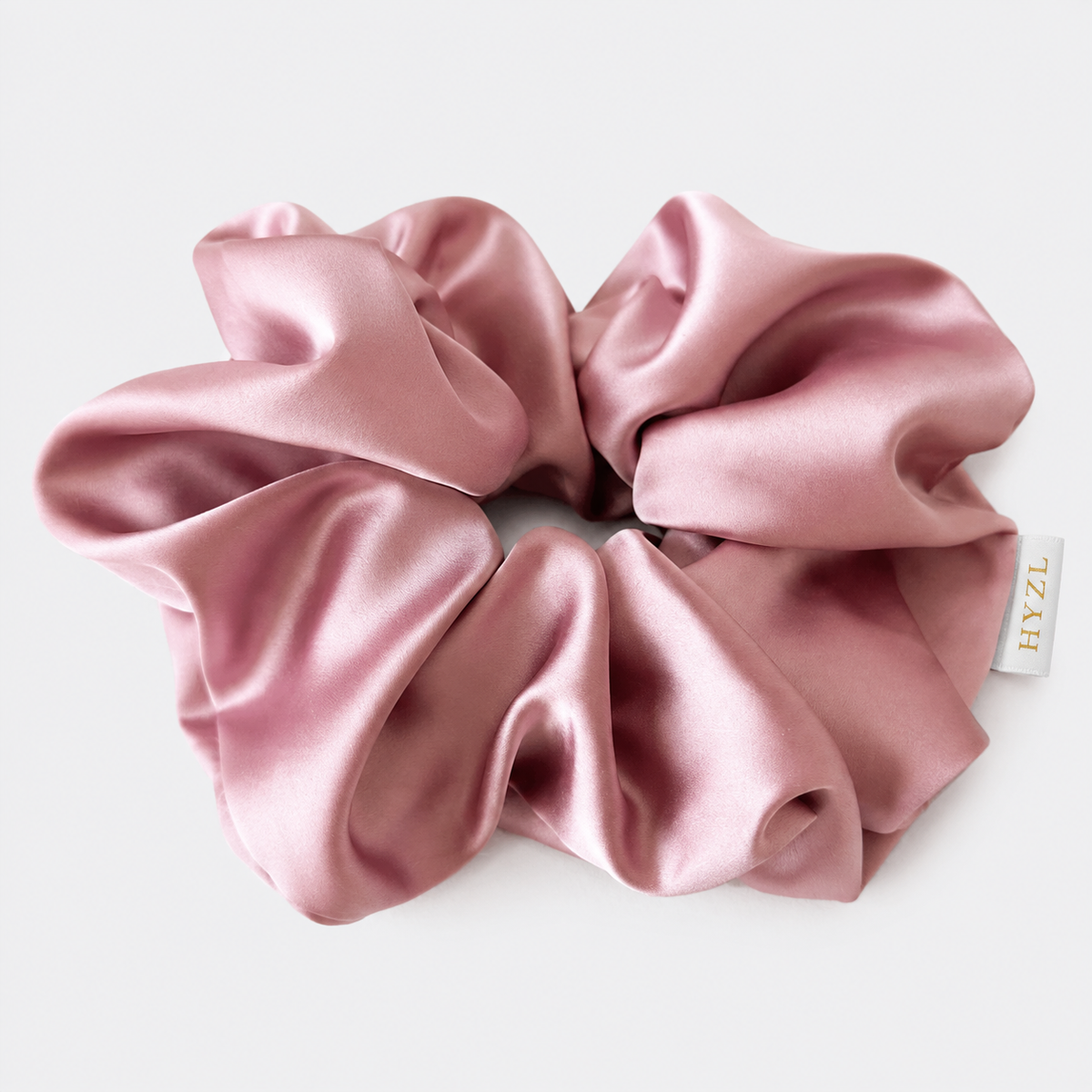 A dusty pink satin hair scrunchie with a soft, lustrous sheen, shown on a clean white background with a small brand tag reading 'HYZL'.