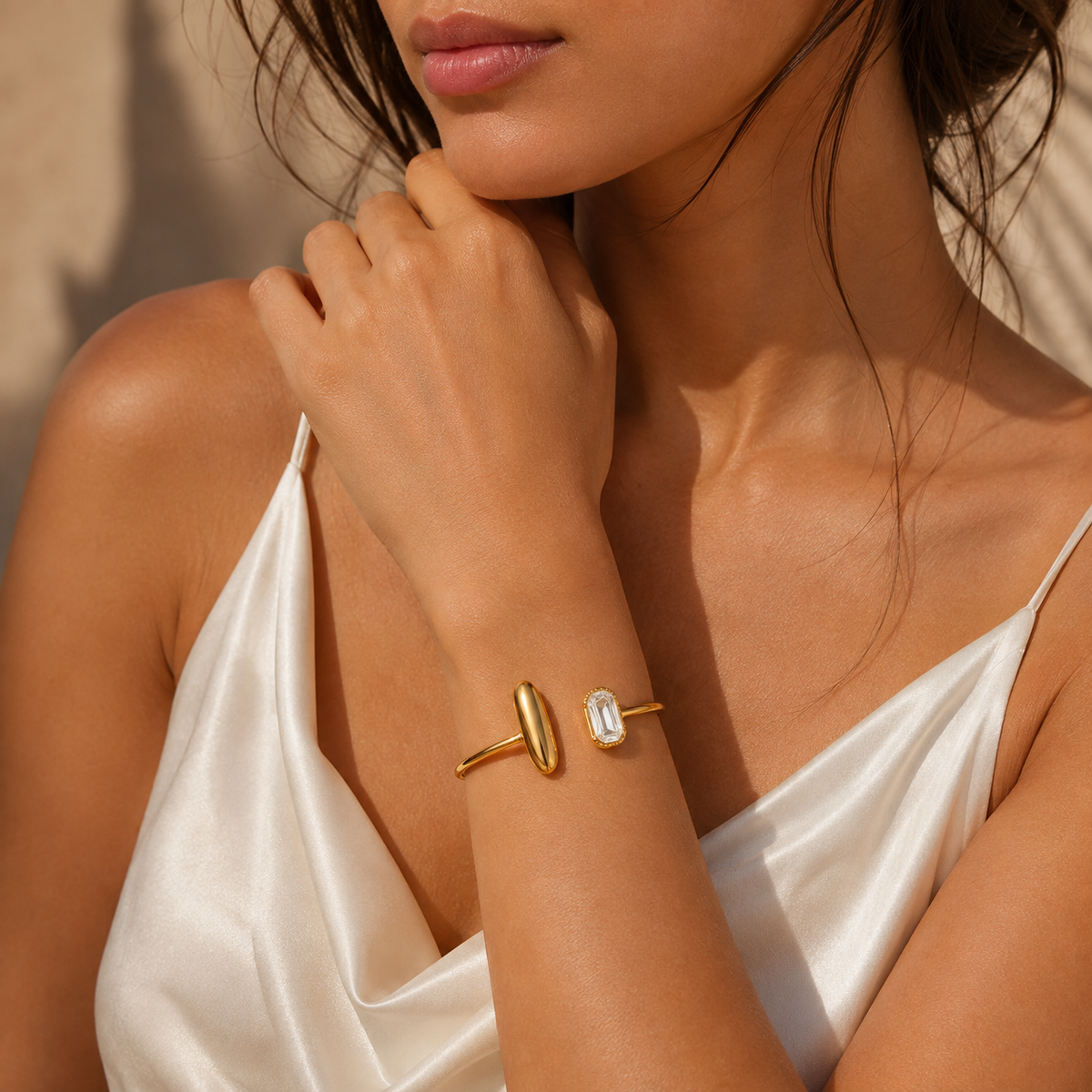 A close-up lifestyle shot of a model wearing a gold cuff bracelet with a vertical oval bar and a second gold cuff featuring a rectangular clear gemstone, set against a warm beige background.