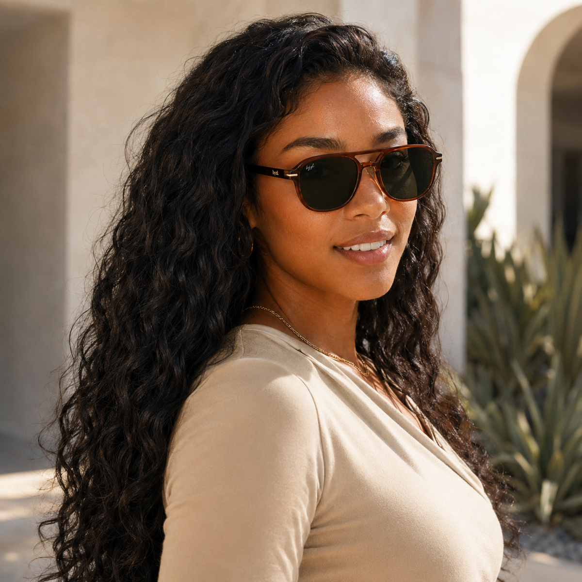A woman with long, curly black hair wearing brown aviator-style sunglasses with dark lenses and a beige top, photographed outdoors against a sunlit architectural background.