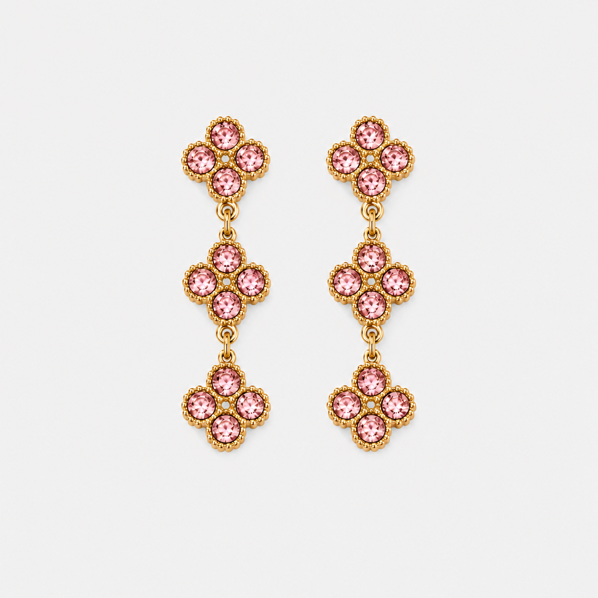 A pair of gold dangle earrings featuring three stacked floral clusters, each set with pink gemstones in a textured gold bezel setting, shown against a light gray background.