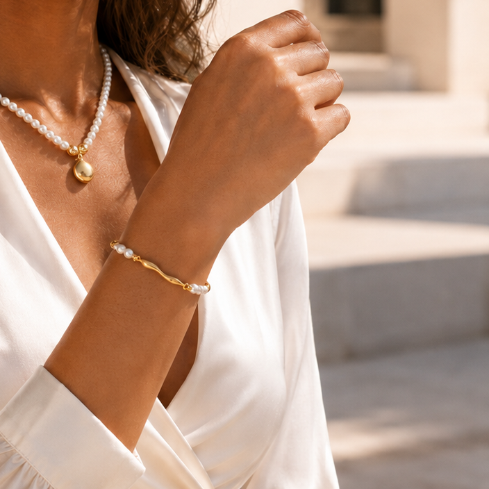 A close-up lifestyle shot of a woman wearing a delicate gold bracelet with white pearl accents on her wrist, paired with a matching pearl necklace, set against a soft-focus outdoor background.