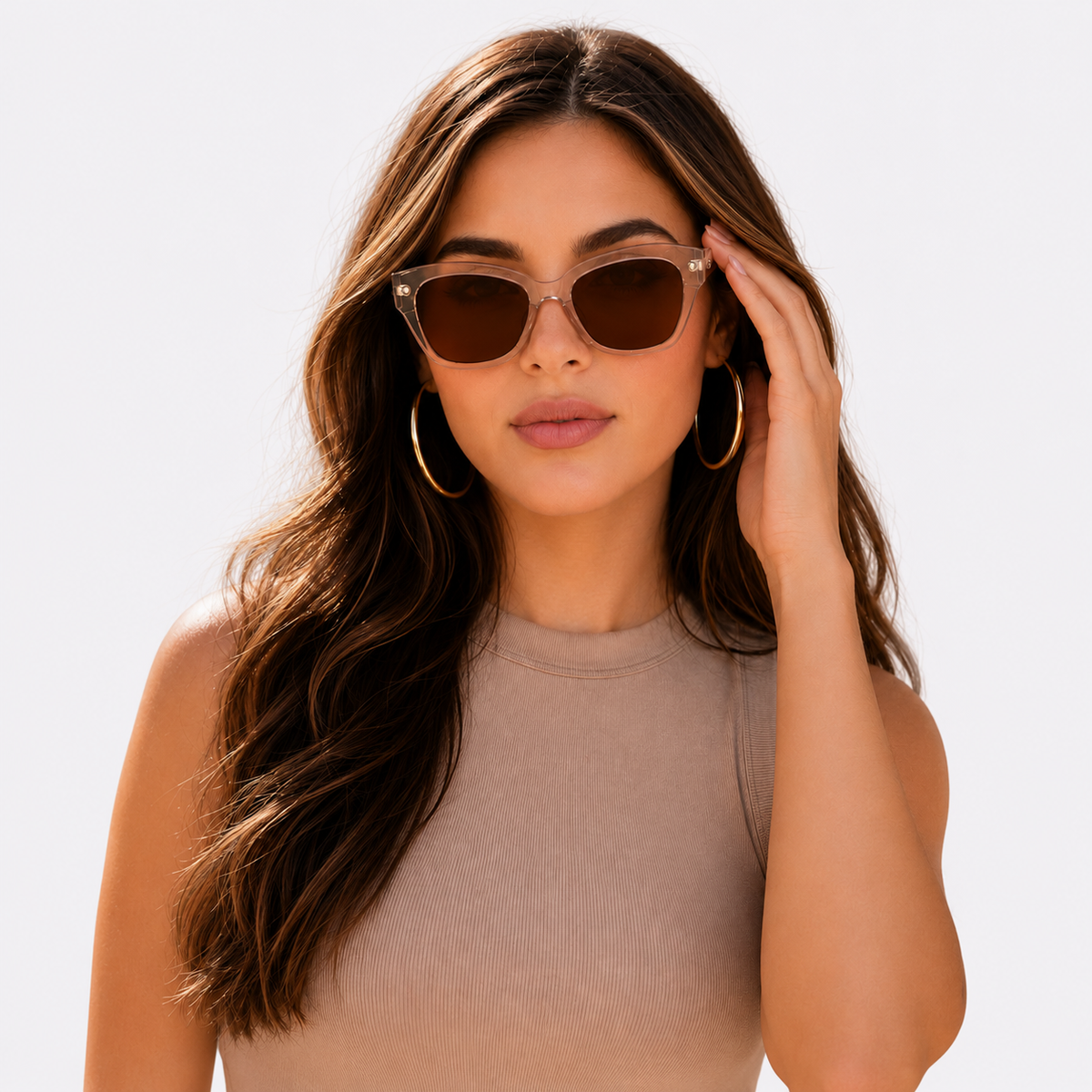 A woman with long wavy brown hair wearing cat-eye sunglasses with clear frames and dark brown lenses, gold hoop earrings, and a beige ribbed top against a light gray background.