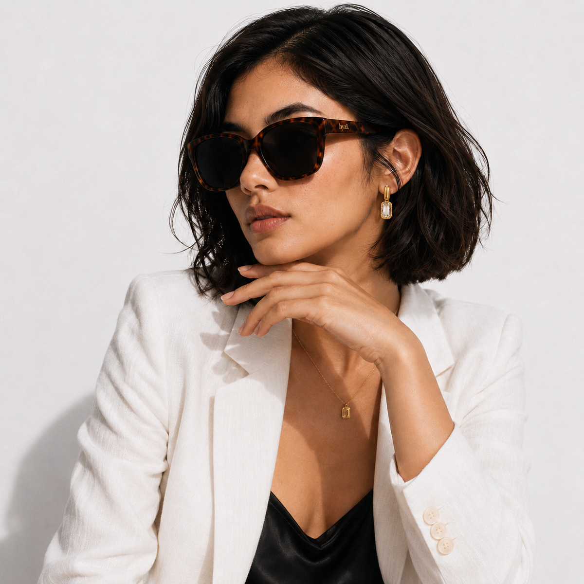 A woman with short dark hair wears tortoiseshell cat-eye sunglasses, a white blazer, and a black top against a light gray background.