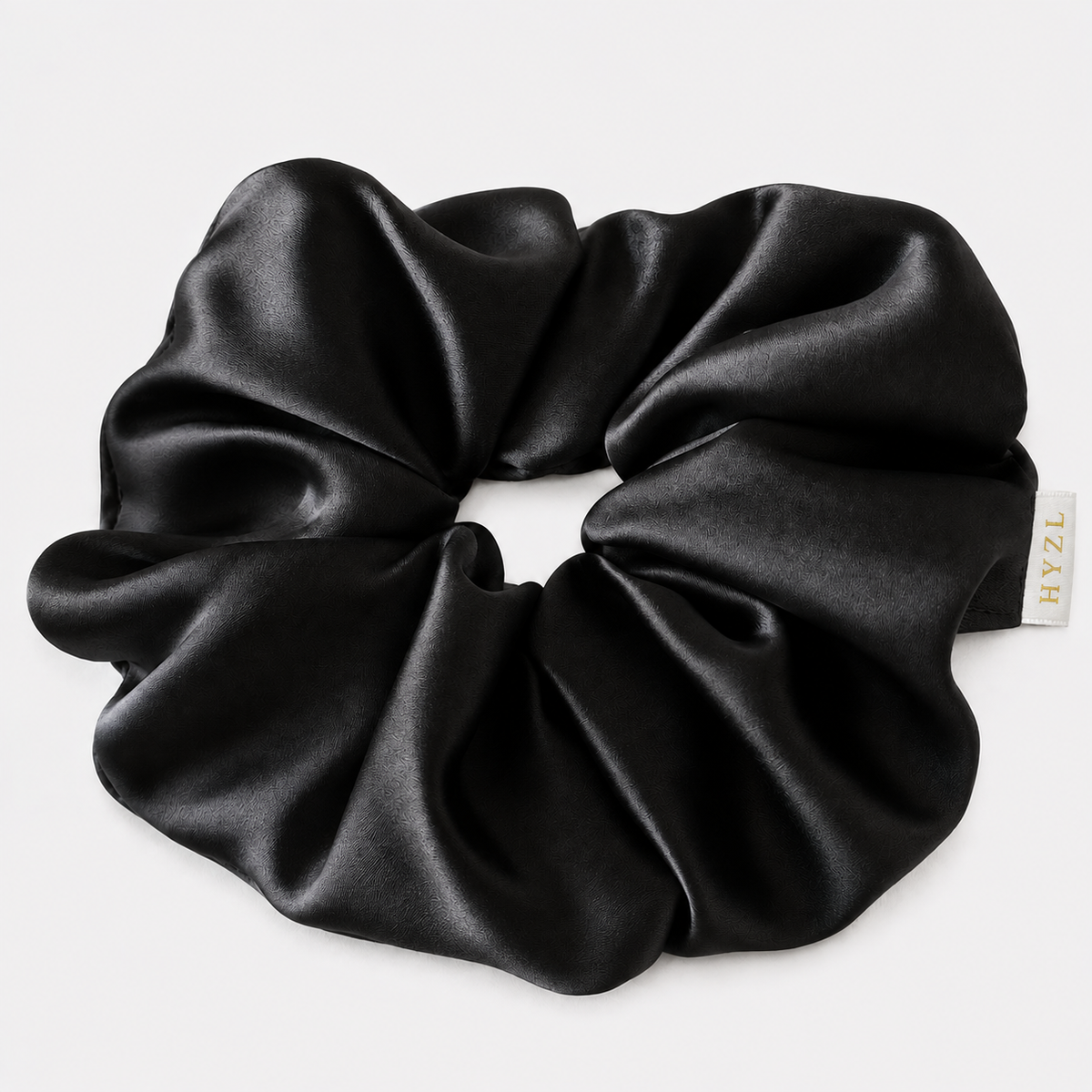 A black satin hair scrunchie with a small white brand tag reading 'HYZL', shown on a clean light gray background.