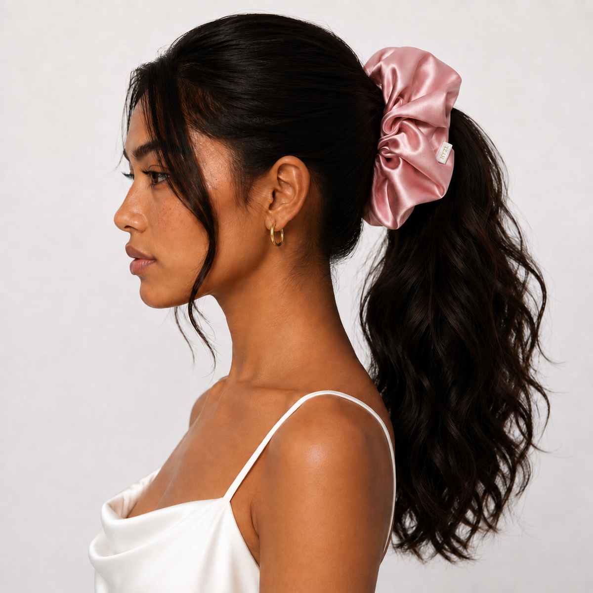 A woman in profile wearing a large, shiny dusty pink satin scrunchie in her ponytail, paired with a white spaghetti-strap top against a light gray background.