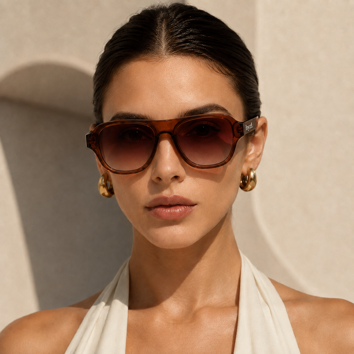 A close-up of a woman wearing brown acetate square sunglasses with a gradient tint and gold hoop earrings, set against a warm beige textured wall.