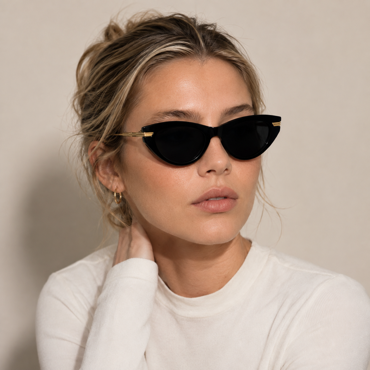 A woman with blonde hair in a messy bun wearing black cat-eye sunglasses with gold metal temples and a white long-sleeve top, photographed against a neutral beige background.
