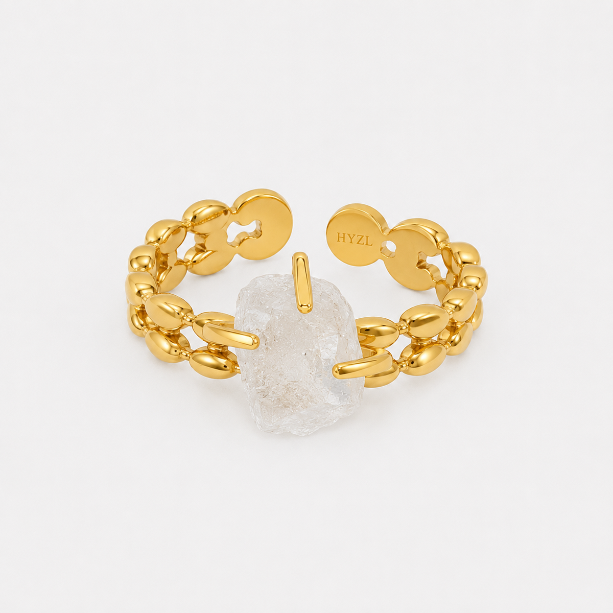 A gold chain-link ring with a central clear quartz crystal, photographed on a clean white background. The inside of the ring is visible, showing a circular tag engraved with the brand name 'HYZL'.