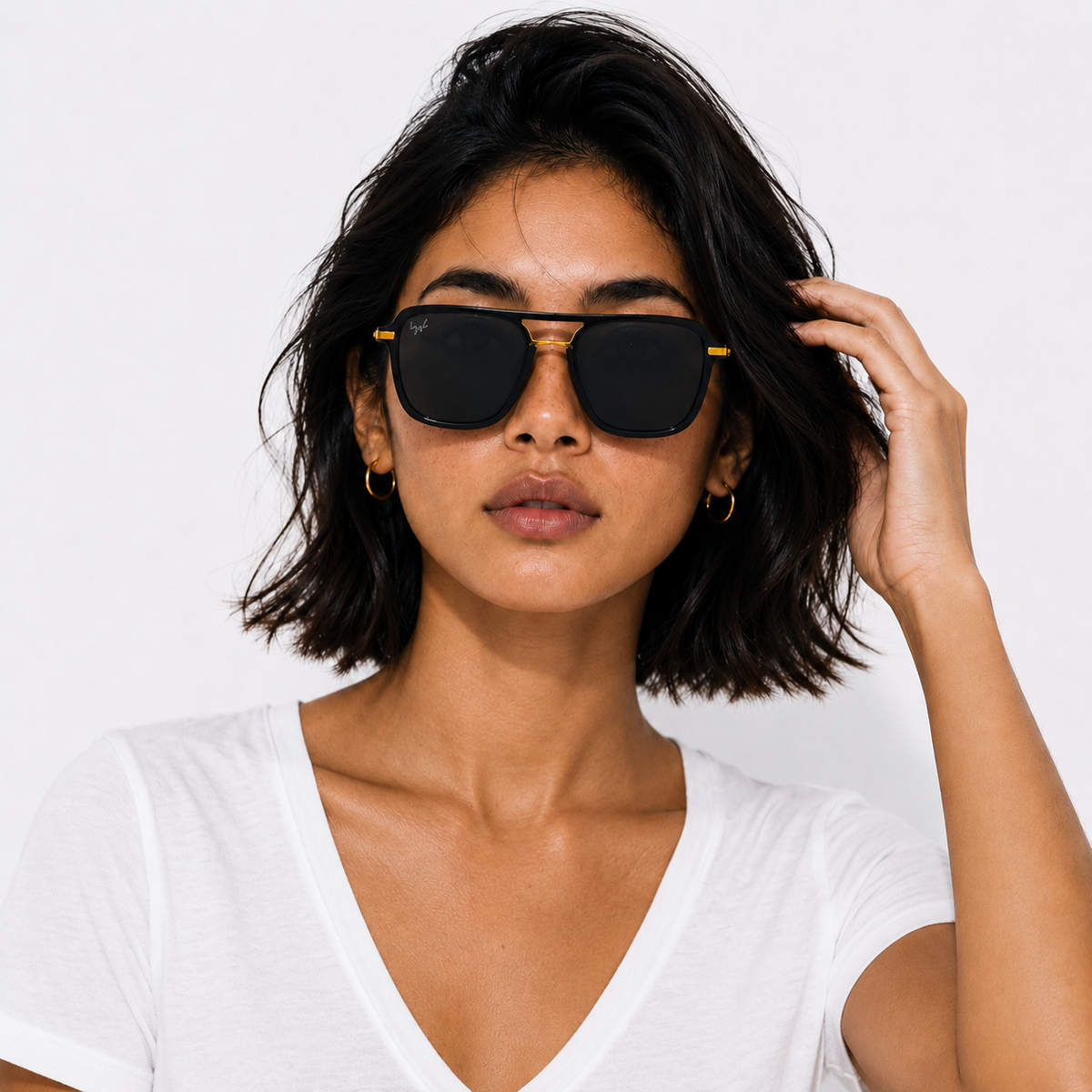 A woman with short dark hair wearing black square sunglasses with gold metal frames and a white V-neck t-shirt, photographed against a clean white background.